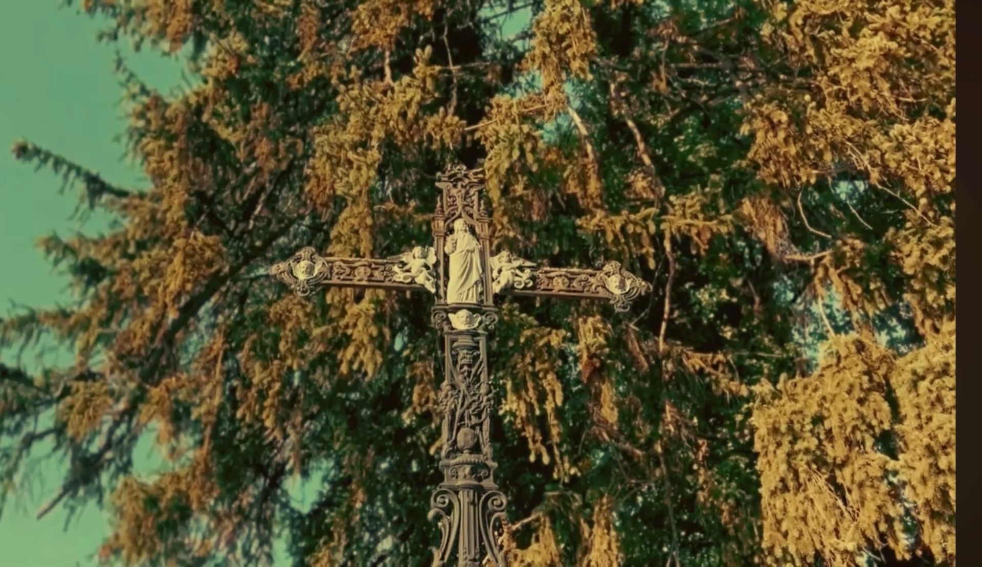 Croix de mission ouvragée portant une statue, entourée de branches de mélèze