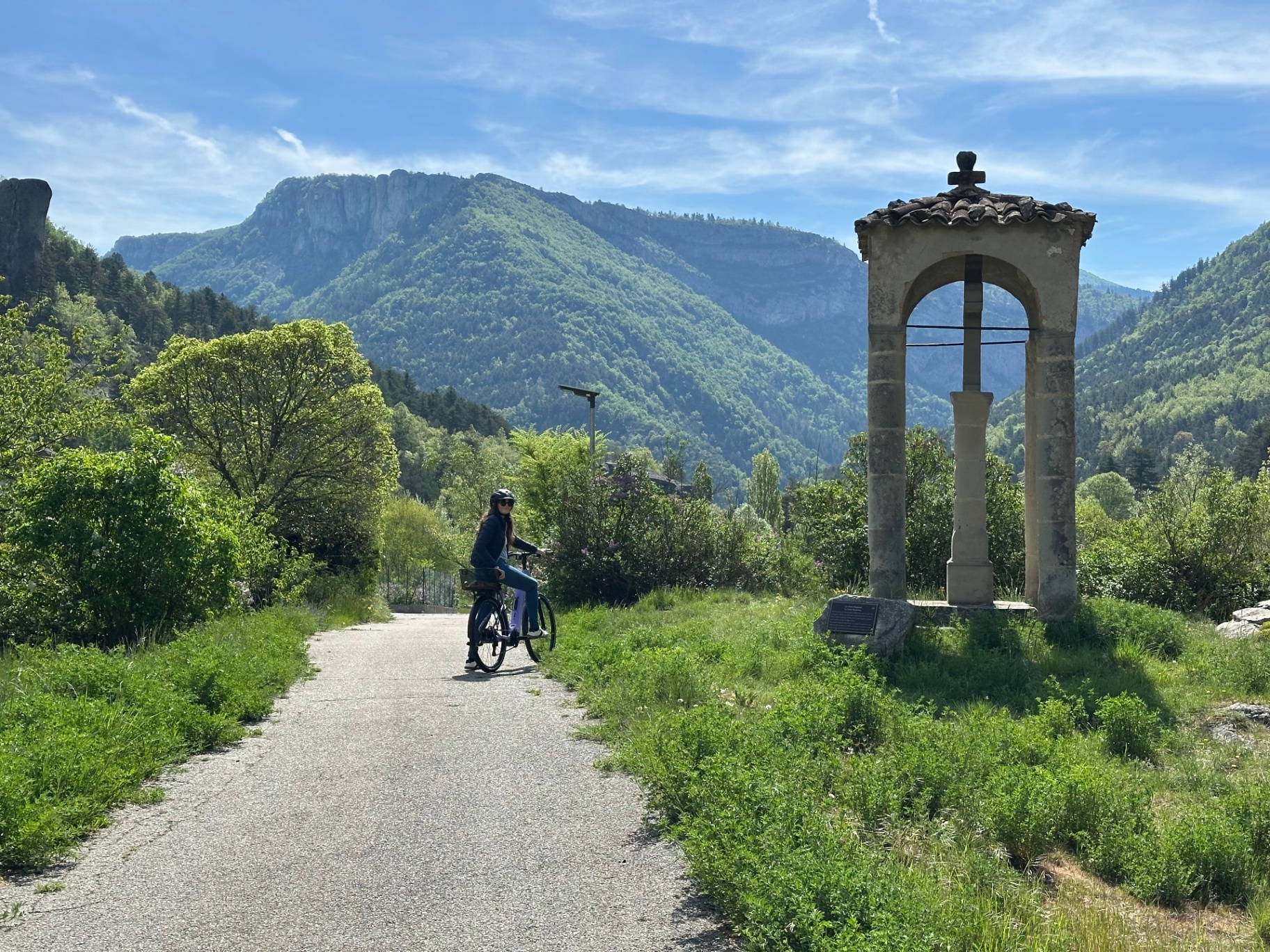 La Croix romane d'Annot au bord du chemin, une cycliste passant devant, les montagnes du pays d'Annot en arrière-plan